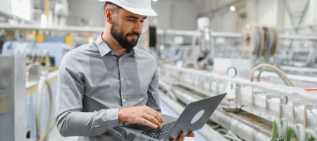 Mann mit Schutzhelm arbeitet an einem Laptop in einer modernen Industrieanlage mit Maschinen und Rohrleitungen im Hintergrund.