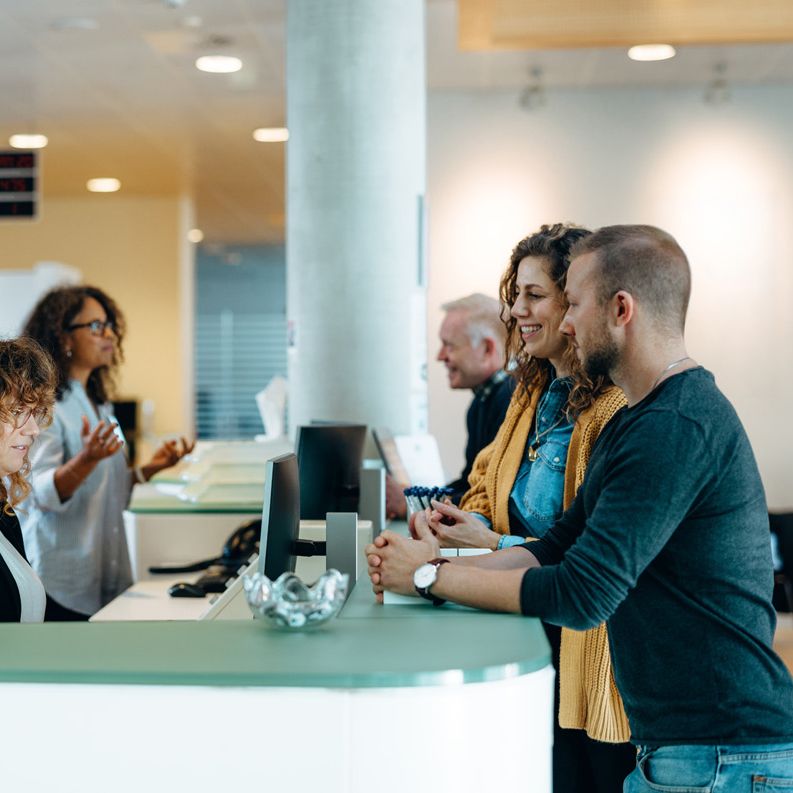 Ein mittelgroßer Mann und eine Frau mit lockigen Haaren stehen an einem geschwungenen Empfangstresen und sprechen mit einer Empfangsmitarbeiterin mit Brille. Die Mitarbeiterin sitzt hinter einem Monitor. Im Hintergrund sind weitere Mitarbeiter und Gäste s