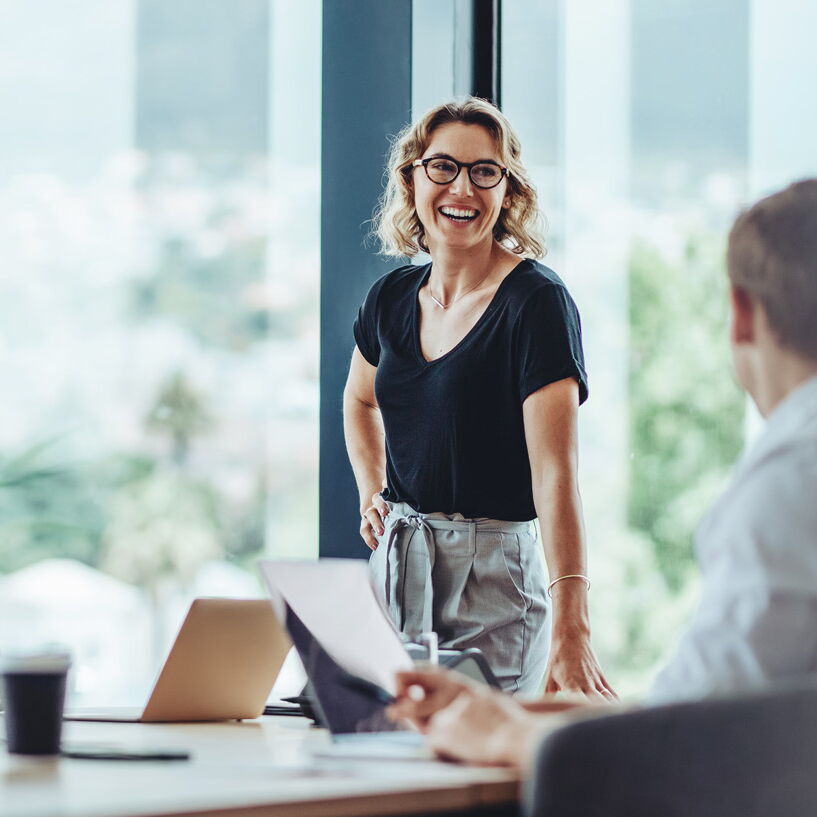 Vier Personen sitzen in einem modernen Büro am Tisch, während eine Frau lächelnd präsentiert und das Team gemeinsam an digitalen Lösungen und Prozessen arbeitet