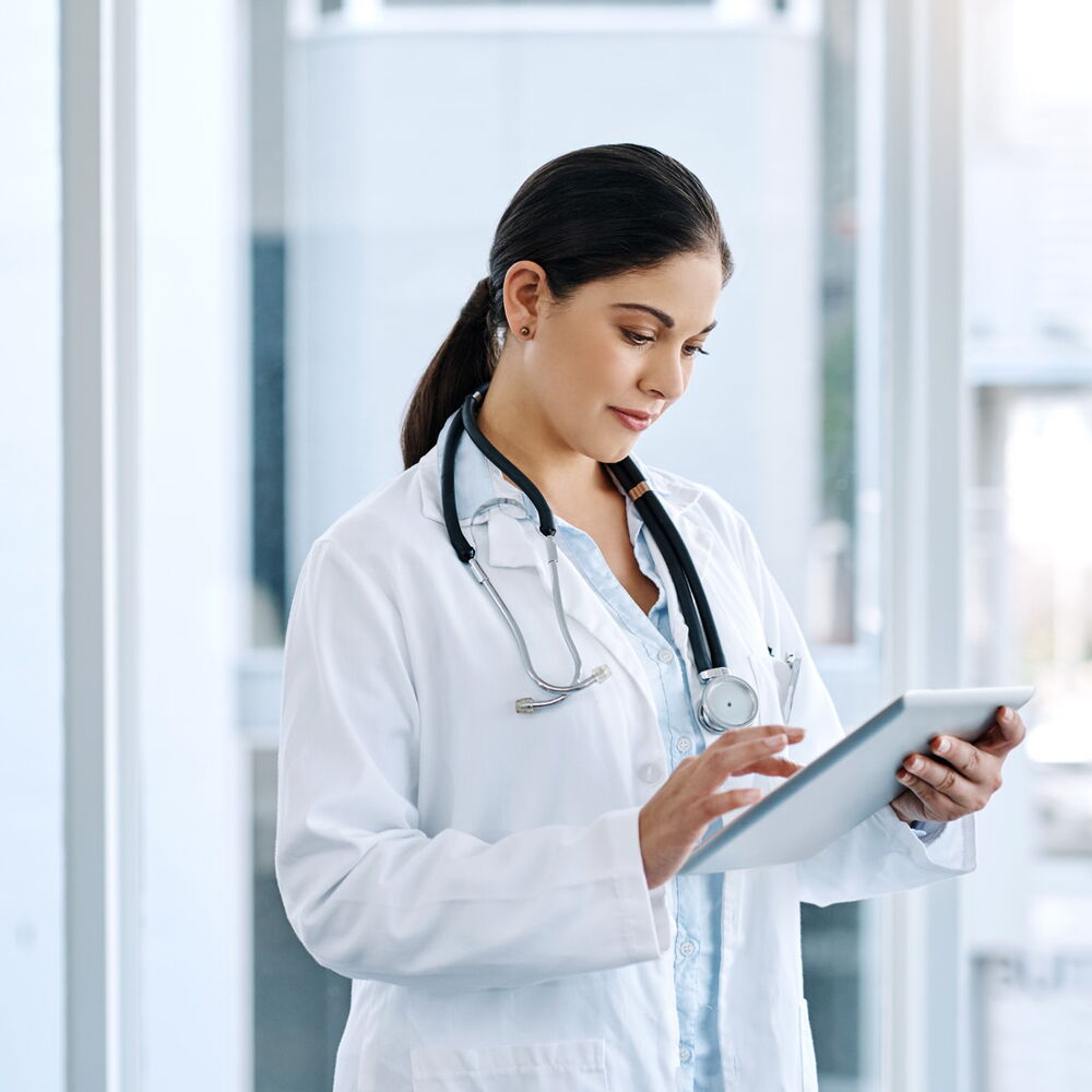 Ärztin im weißen Kittel mit Stethoskop nutzt ein Tablet in einer hellen Klinik und arbeitet digital mit Patientendaten und Prozessen