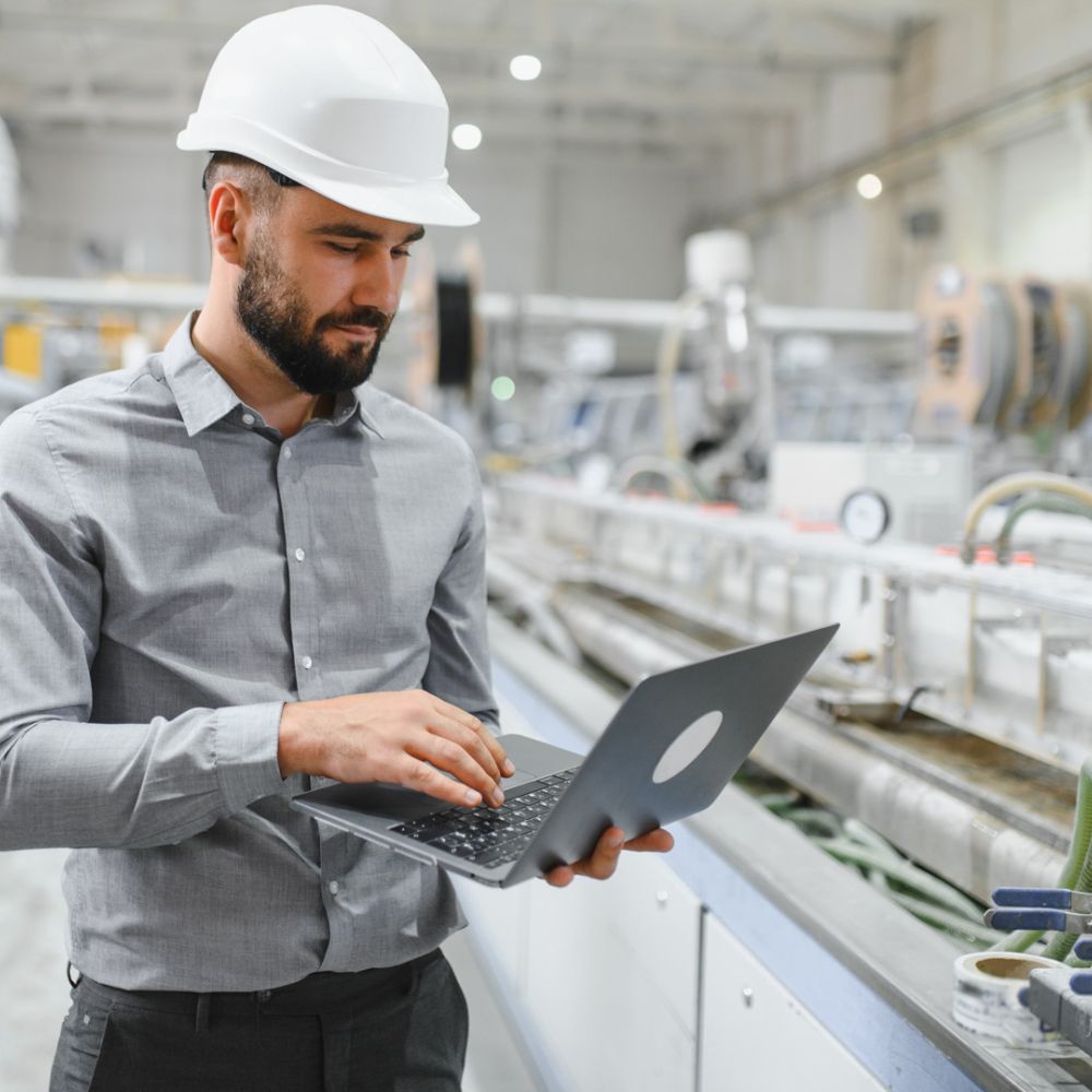 Mann mit Schutzhelm arbeitet an einem Laptop in einer modernen Industrieanlage mit Maschinen und Rohrleitungen im Hintergrund.