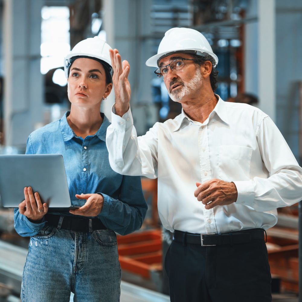 Zwei Ingenieure mit weißen Schutzhelmen in einer Fabrikhalle. Eine Frau in Jeanshemd hält einen Laptop, während ein älterer Mann mit Brille und weißem Hemd auf etwas in der Ferne zeigt und erklärt.
