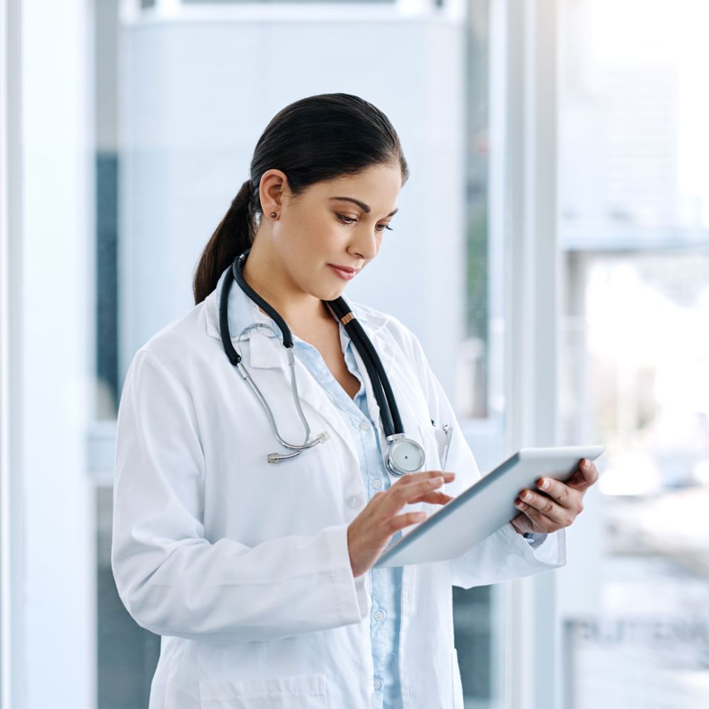 Hier ist ein passender Alt-Text für das Bild:  Alt-Text: Eine junge Ärztin mit dunklen, zum Zopf gebundenen Haaren steht in einem hellen, modernen Krankenhausflur und bedient ein Tablet. Sie trägt einen weißen Laborkittel über einem blauen Hemd und ein St