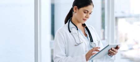 Hier ist ein passender Alt-Text für das Bild:  Alt-Text: Eine junge Ärztin mit dunklen, zum Zopf gebundenen Haaren steht in einem hellen, modernen Krankenhausflur und bedient ein Tablet. Sie trägt einen weißen Laborkittel über einem blauen Hemd und ein St