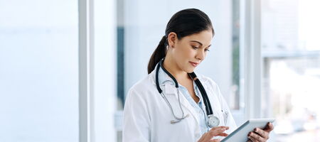 Ärztin im weißen Kittel mit Stethoskop nutzt ein Tablet in einer hellen Klinik und arbeitet digital mit Patientendaten und Prozessen