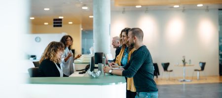 Ein mittelgroßer Mann und eine Frau mit lockigen Haaren stehen an einem geschwungenen Empfangstresen und sprechen mit einer Empfangsmitarbeiterin mit Brille. Die Mitarbeiterin sitzt hinter einem Monitor. Im Hintergrund sind weitere Mitarbeiter und Gäste s