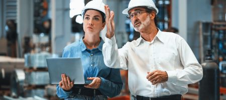 Zwei Ingenieure mit weißen Schutzhelmen in einer Fabrikhalle. Eine Frau in Jeanshemd hält einen Laptop, während ein älterer Mann mit Brille und weißem Hemd auf etwas in der Ferne zeigt und erklärt.