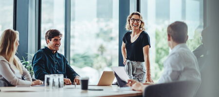 Vier Personen sitzen in einem modernen Büro am Tisch, während eine Frau lächelnd präsentiert und das Team gemeinsam an digitalen Lösungen und Prozessen arbeitet